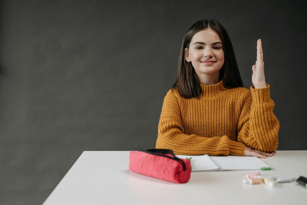 A cheerful young girl raising her hand in a classroom setting, ready to answer.